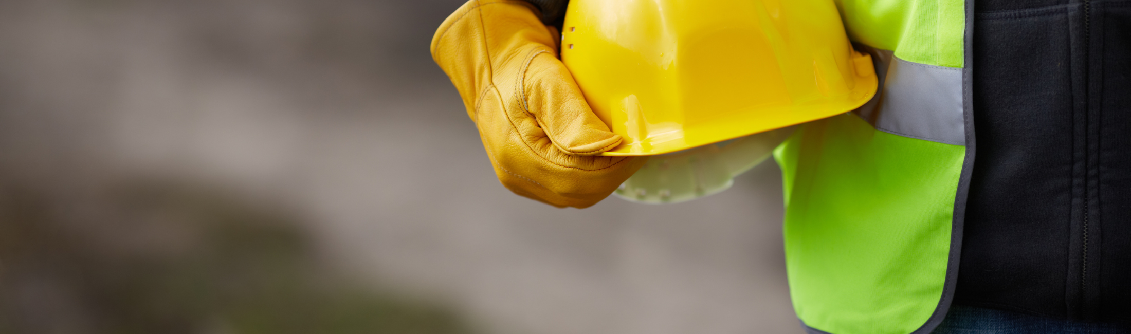 person holding hard hat at hip wearing leather construction gloves and safety vest