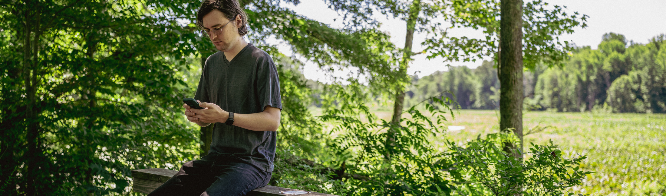 Man with glasses sitting on bench in the woods looking at a smart phone